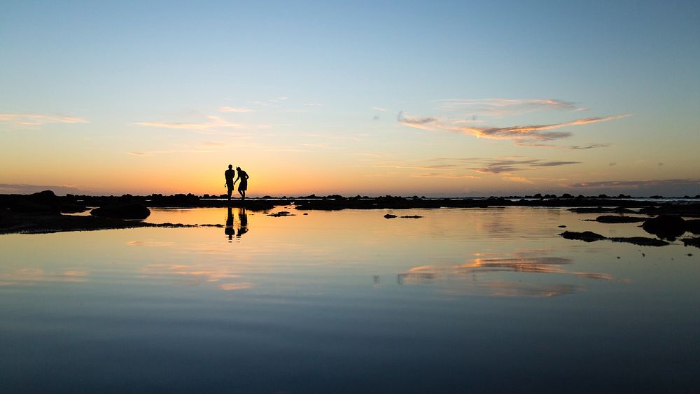 A silhouetted couple stand together on the horizon as they and the blue and yellow sky reflect into the still water…