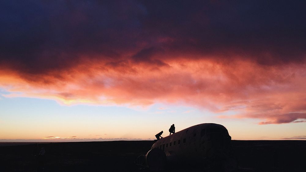 Two people climbing on an abandoned airplane under a sunset sky with pink clouds in Solheimasandur. Original public domain…
