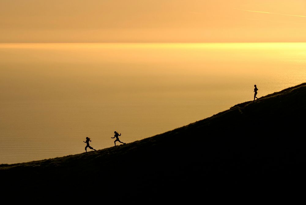 Three women energetically running down Mount Tamalpais as waters in the distance reflect the golden sunset. Original public…