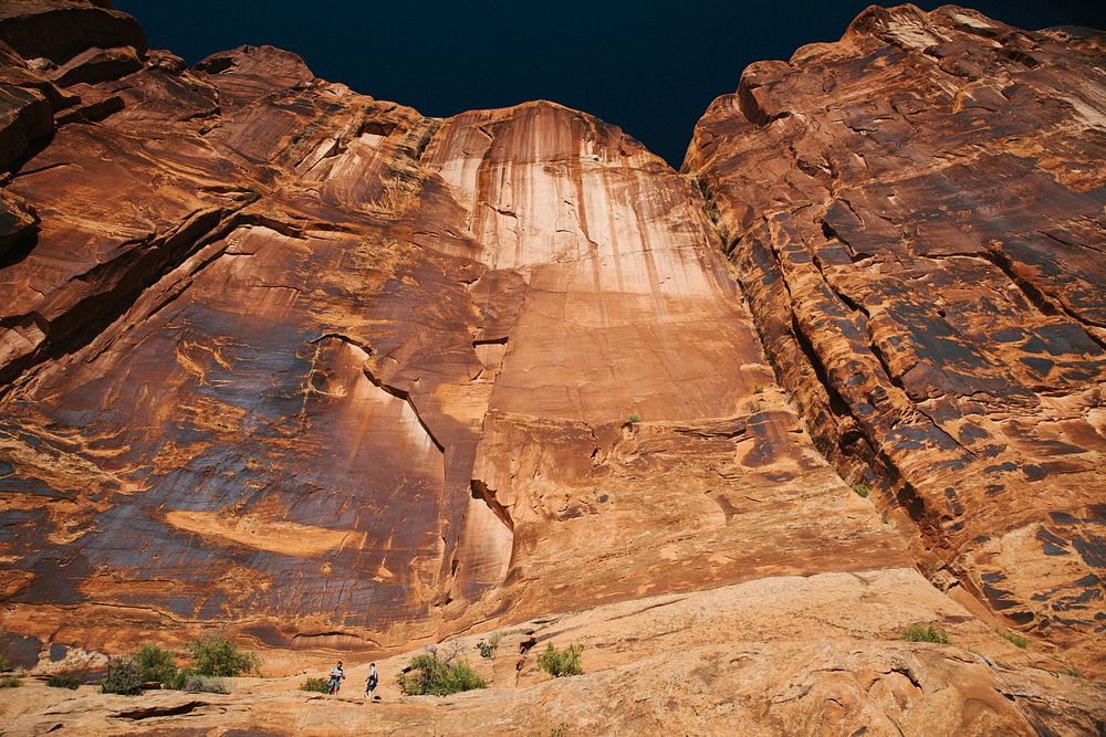 Two people at the foot of a tall vertical face of red rock. Original public domain image from Wikimedia Commons