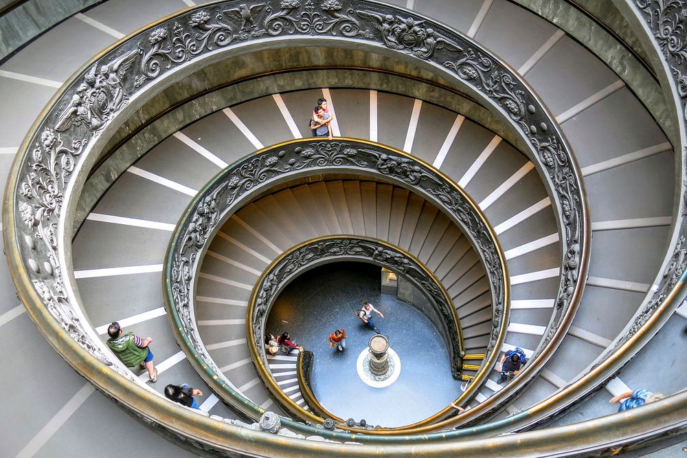 Looking down a massive spiral staircase while people climb up in the Vatican Museums. Original public domain image from…