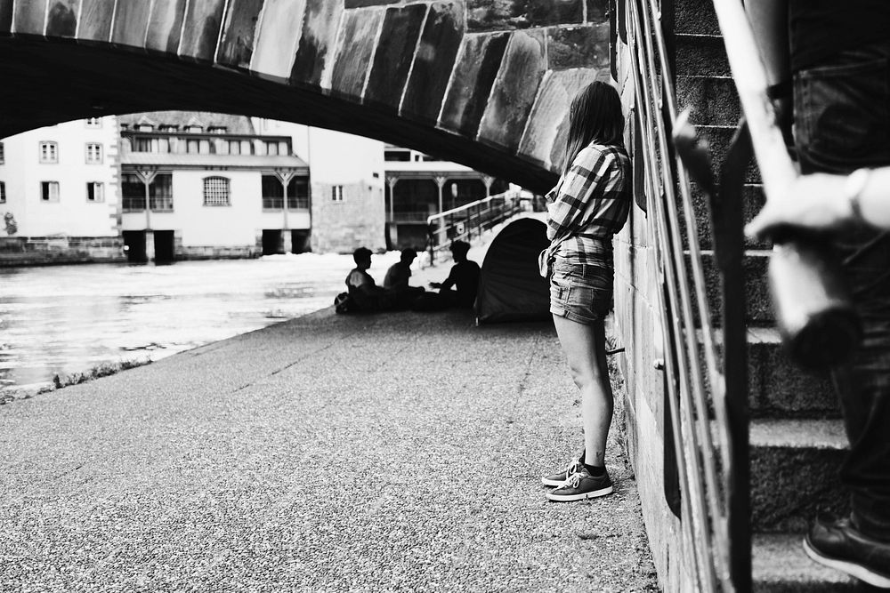 A black-and-white shot of a girl standing near a bridge and looking at people sitting under the bridge. Original public…