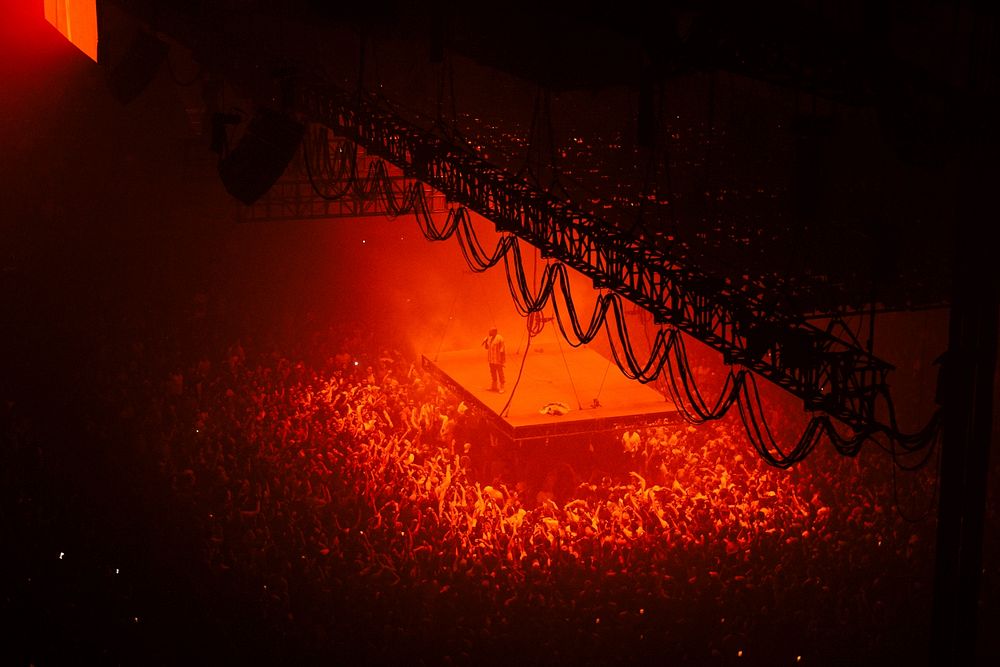 A high shot of a crowd surrounding a suspended stage with a vocalist bathed in red light. Original public domain image from…