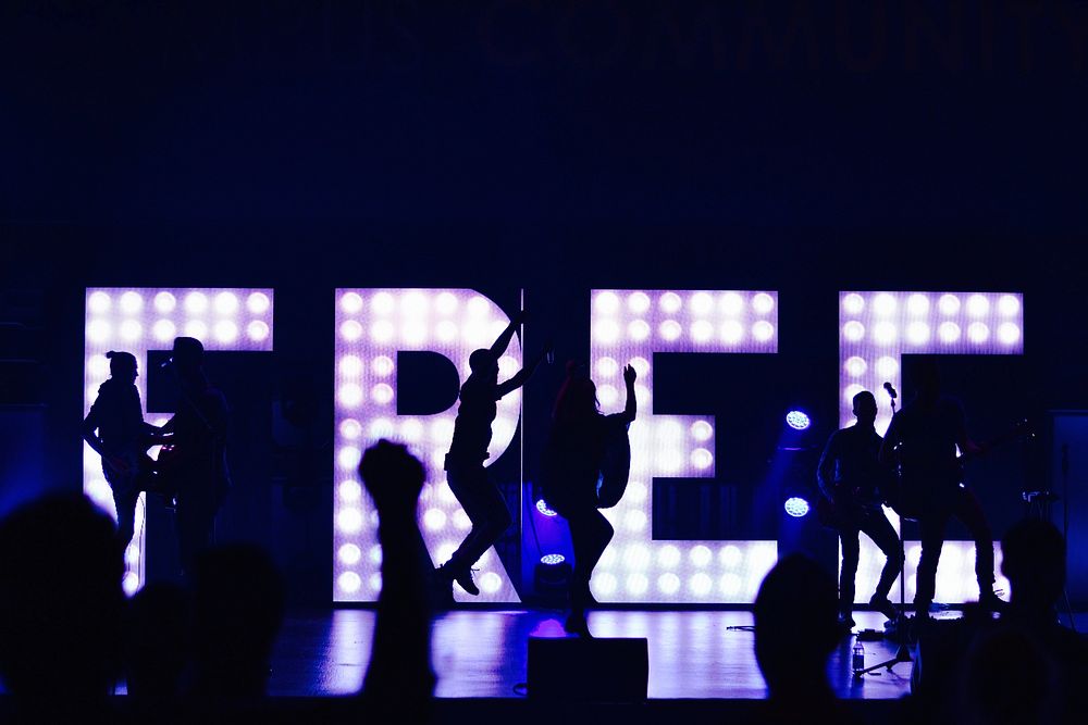 Silhouettes of band members performing and jumping up in front of lights arranged to spell out “free”. Original public…