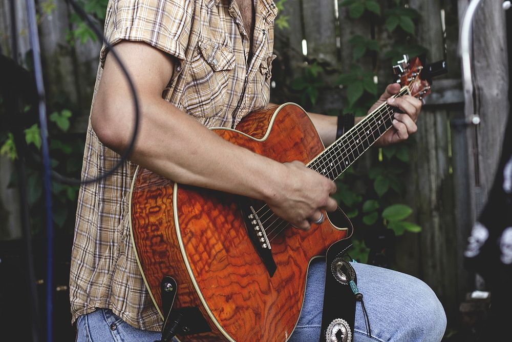 A man in a checkered shirt strumming an acoustic guitar in a garden. Original public domain image from Wikimedia Commons