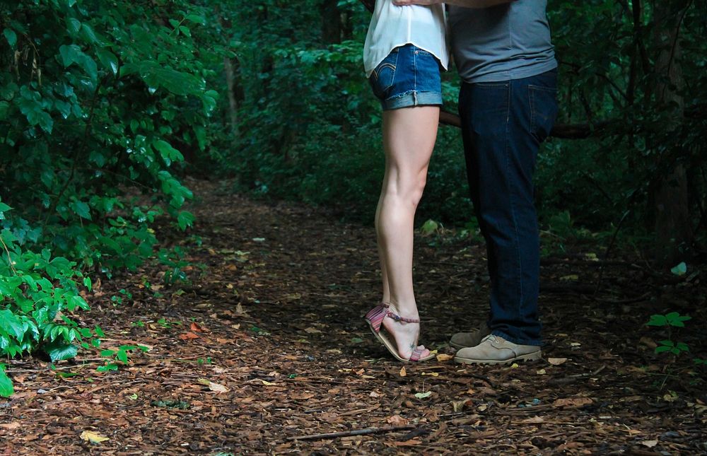 On a forest trail, a woman in flip-flops rises on tip-toes to kiss her man. Original public domain image from Wikimedia…
