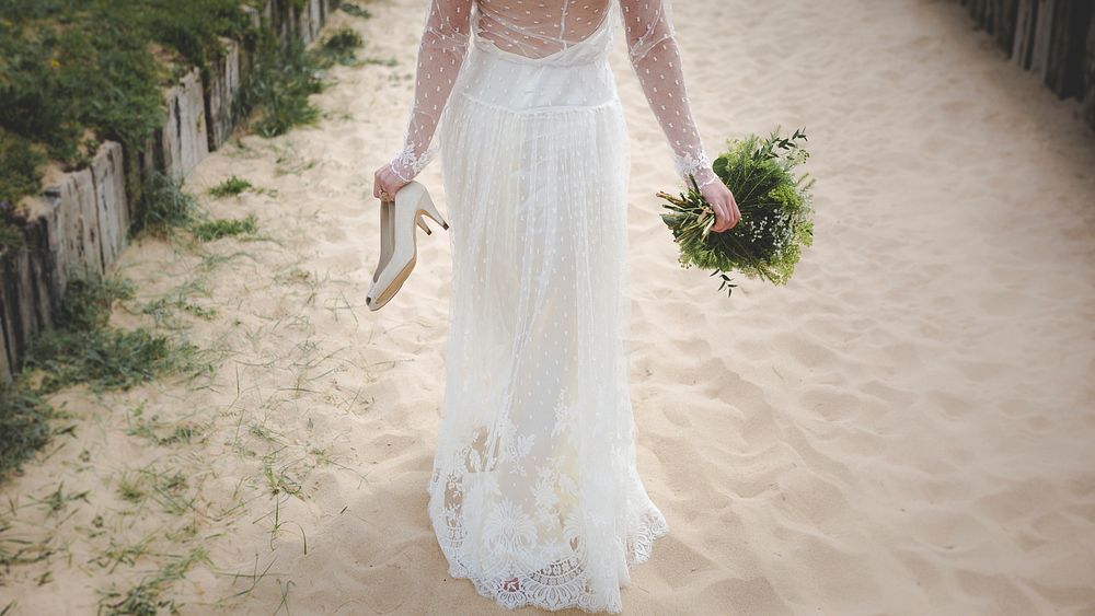 Bride holds shoes and bouquet while walking in sands of Sainte-Marie-de-Ré. Original public domain image from Wikimedia…