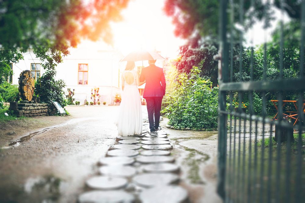 Bride and groom walking down a stone path holding an umbrella with light shining on them. Original public domain image from…