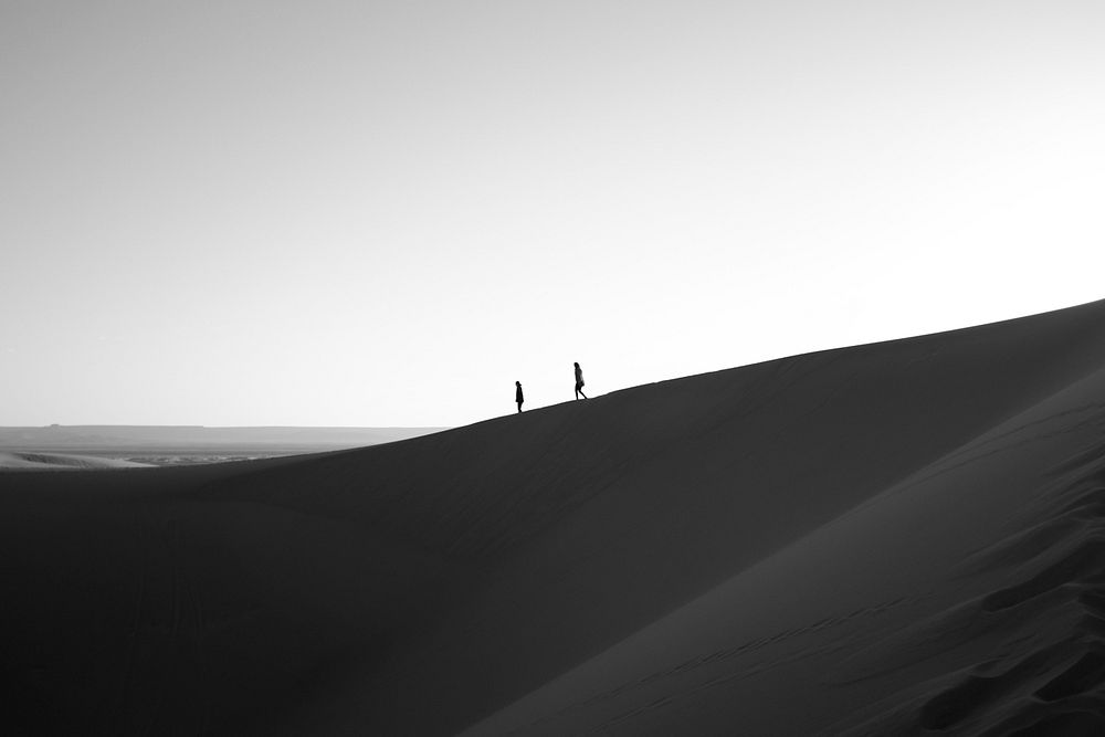 Two people walking across sand dunes in distance, black and white. Original public domain image from Wikimedia Commons