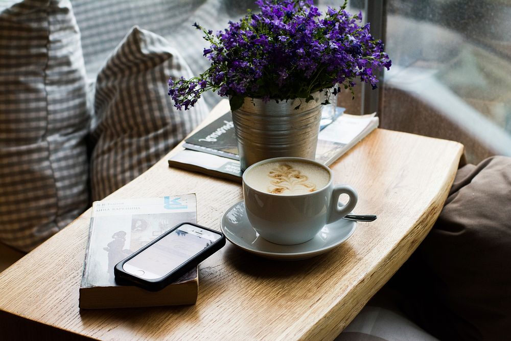 A cup of coffee with latte art on a coffee table next to an iPhone, a purple bouquet and a book. Original public domain…