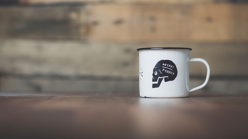 Tin white mug with black skull reading Decaf on it in front of wooden background. Original public domain image from…
