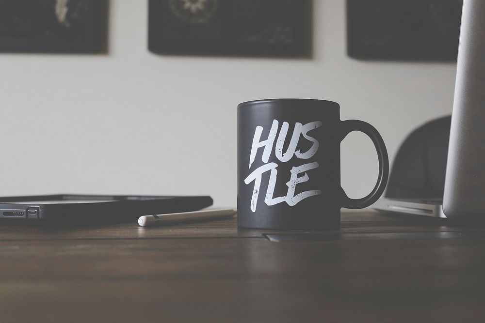 A black mug sitting on an office desk in Missoula with white writing which says "Hustle". Original public domain image from…