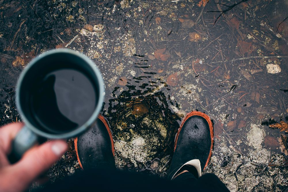 A person wearing rain boots stands in a puddle looking down and holding coffee. Original public domain image from Wikimedia…