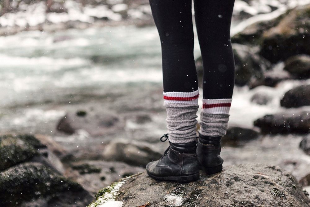 A person with leggings, black boots, and bunched socks stands on a rock during a light snow; only the person's legs are in…