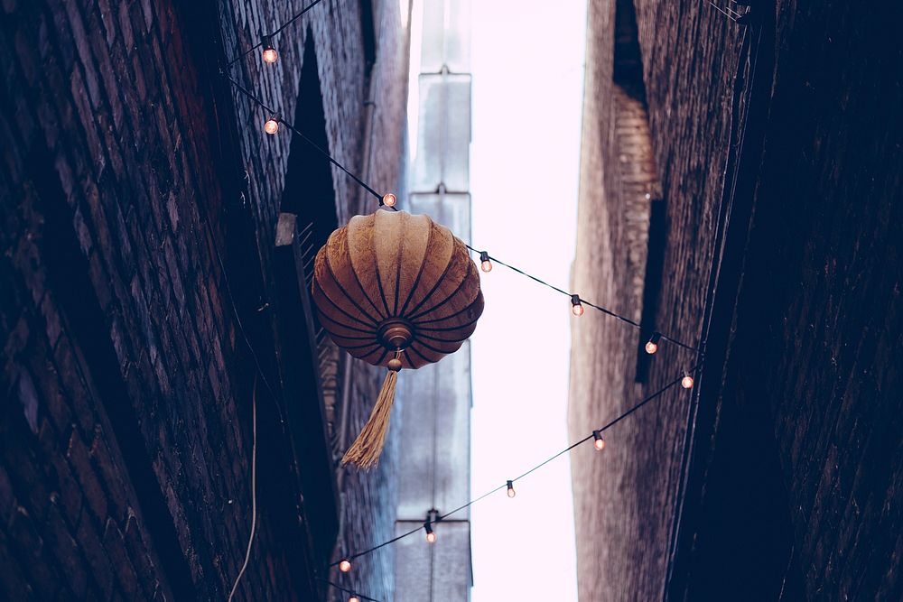Orange Asian lantern hanging from lights seen from the bottom of an alley with tall buildings stretching out of view.…