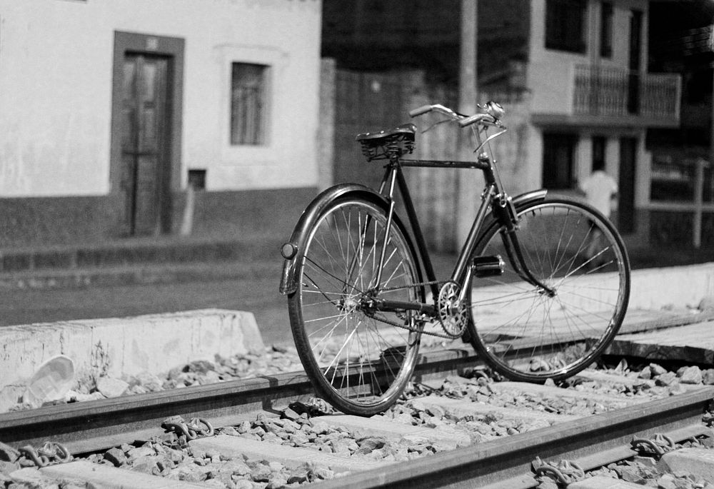 Black and white shot of lone bike standing on railroad tracks with building in background. Original public domain image from…