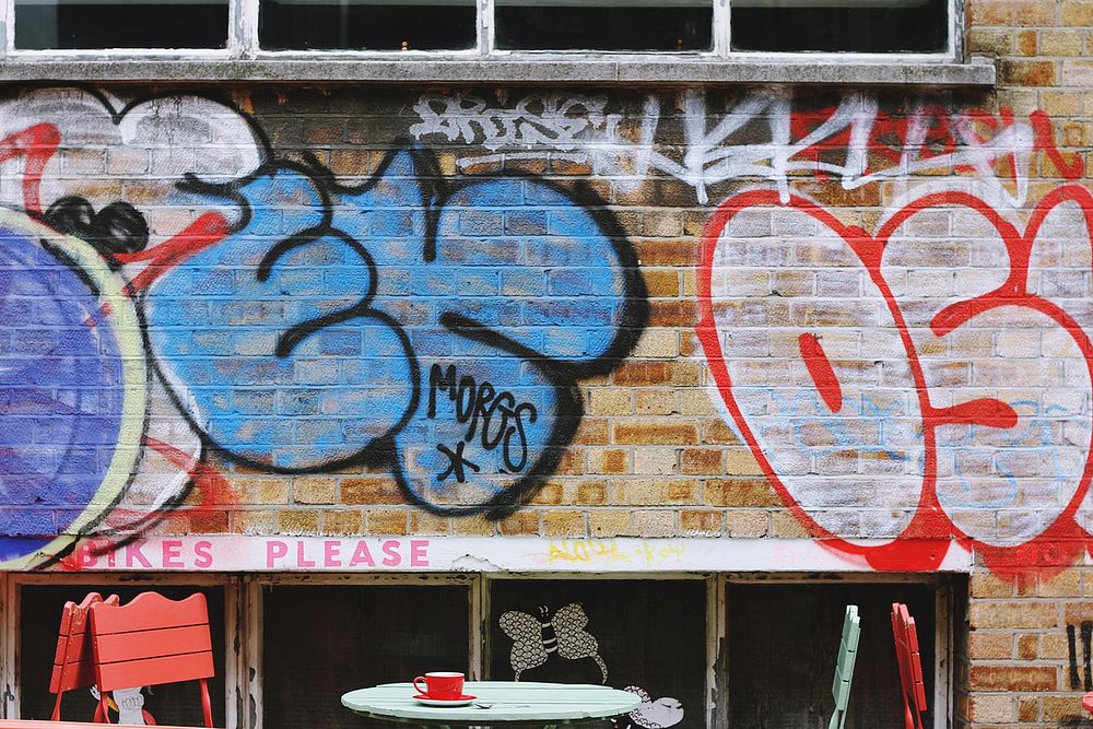 Grunge graffiti on brick wall behind colorful tables and chairs in Shoreditch. Original public domain image from Wikimedia…
