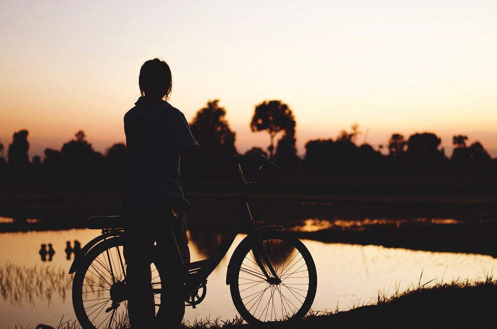 A person in silhouette stands next to a bicycle by a lake at sunset. Original public domain image from Wikimedia Commons