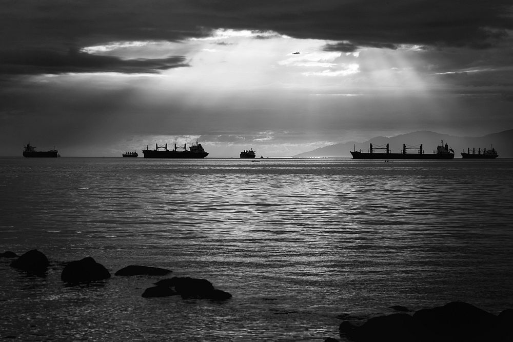 A black and white photo of boats on a calm sea on a beautiful day with light shining through the clouds. Original public…
