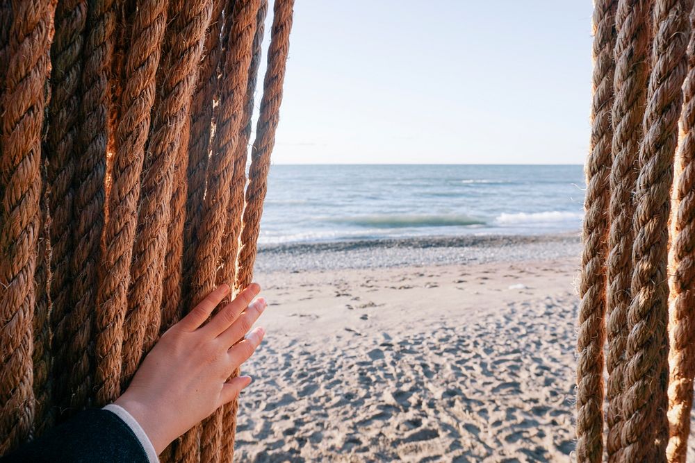 A person's hands pushing long hanging ropes aside on a sandy beach. Original public domain image from Wikimedia Commons