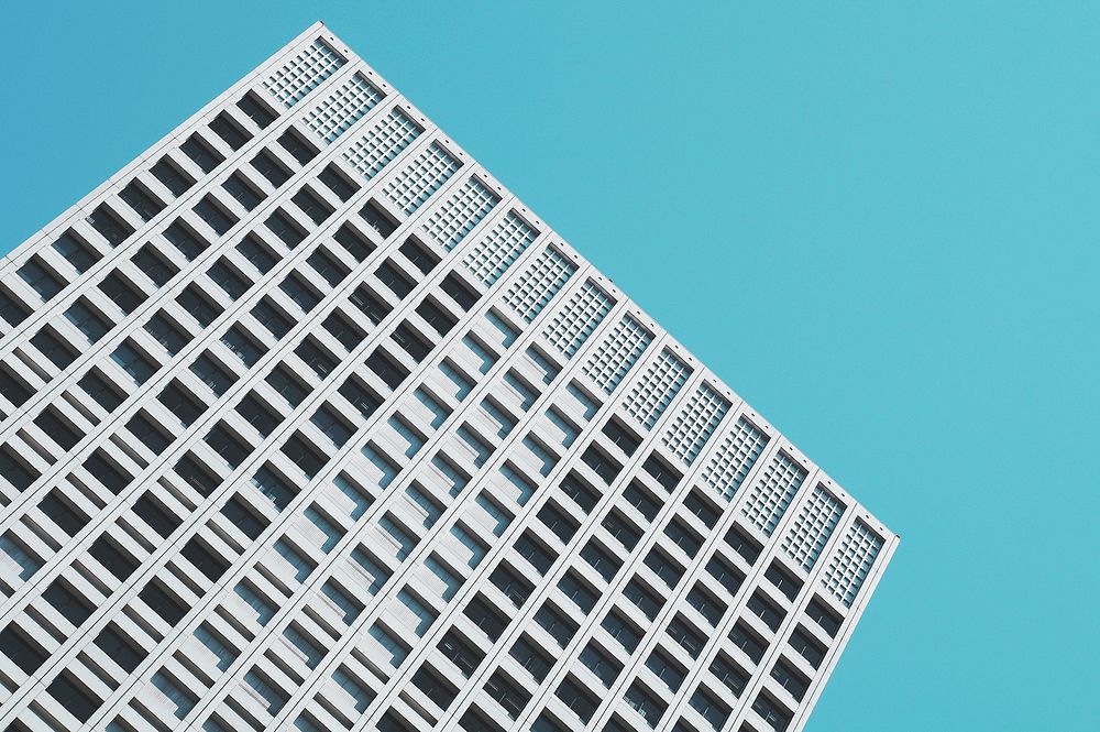 A minimalist concrete skyscraper building with many windows stands at an angle under blue sky. Original public domain image…