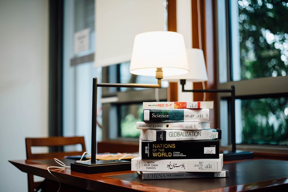 Interior with lamps on a wooden table and a stack of books near a window. Original public domain image from Wikimedia Commons