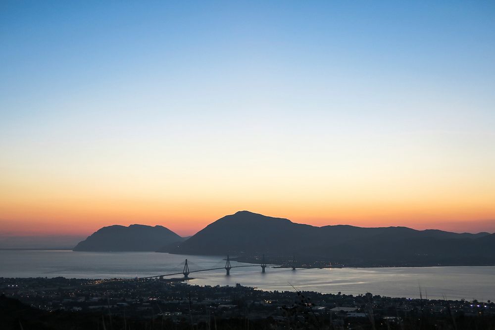 A sunset over a distant mountain and bridges over the river, with the city of Patras in the foreground. Original public…