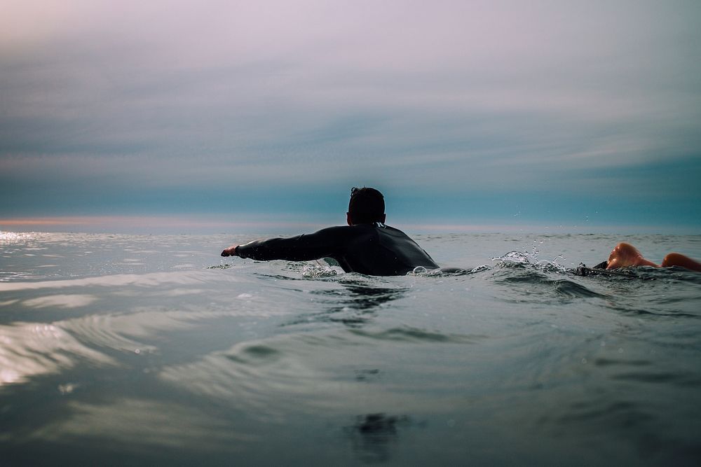 A male surfer swimming in a calm sea under a cloudy sky in Raglan. Original public domain image from Wikimedia Commons