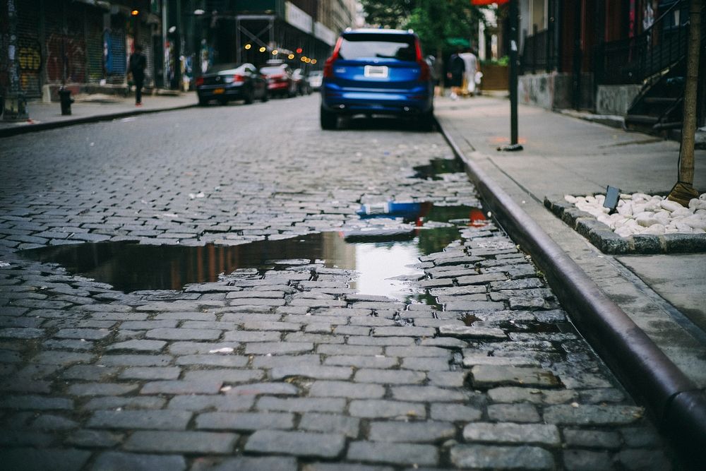 Cobblestone street with parked car and puddle reflecting sky. Original public domain image from Wikimedia Commons