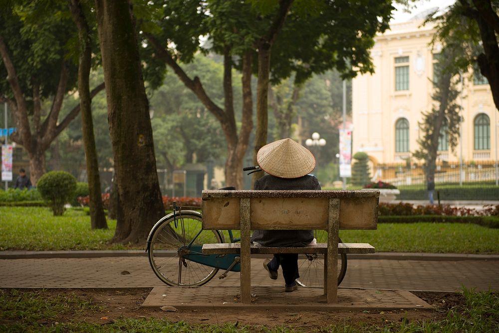 A person wearing a traditional Vietnamese hat sitting on a park bench with their bicycle nearby in Hanoi. Original public…