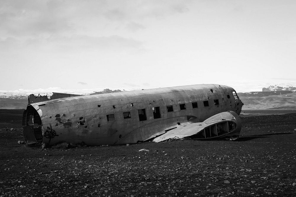 Black and white picture of plane wreck on stony ground with mountains in background, Iceland. Original public domain image…