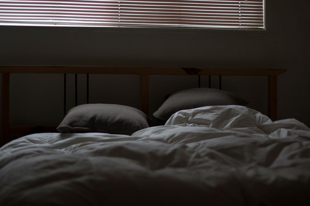 Ruffled gray covers and sheets with two pillows and a large window with blinds above it. Original public domain image from…