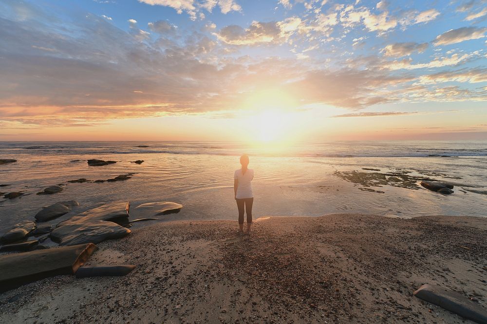 A woman stands on a calm shoreline with peaceful water and a clear sunrise with wispy morning clouds.. Original public…
