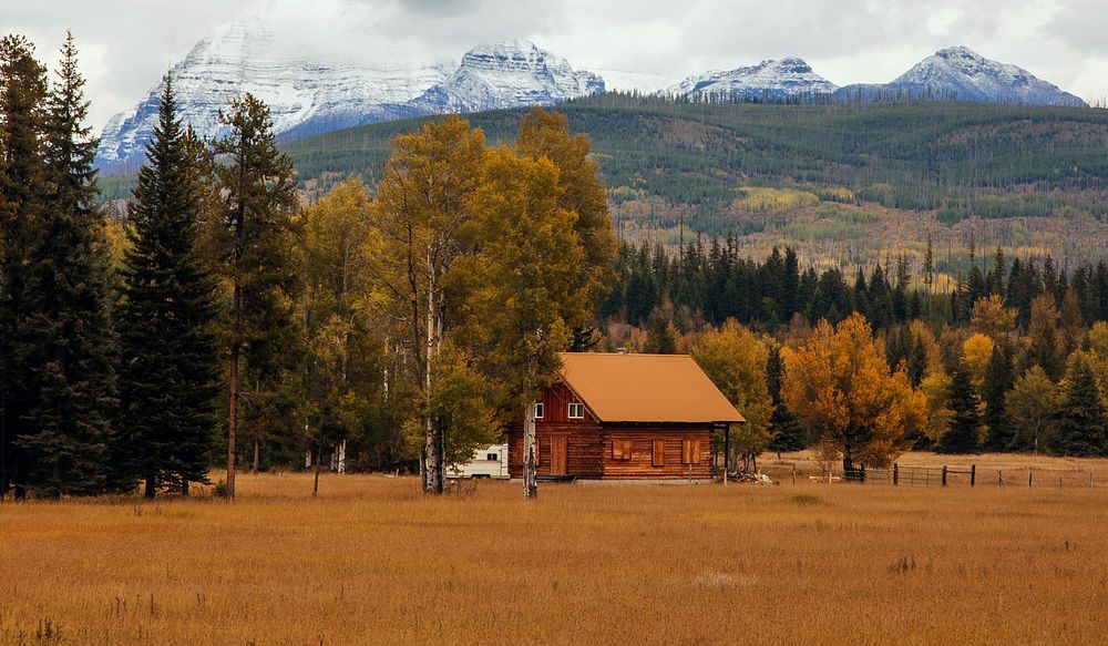 Small log cabin house in Glacier National Park at the edge of a forest and field with snow capped mountains in the…