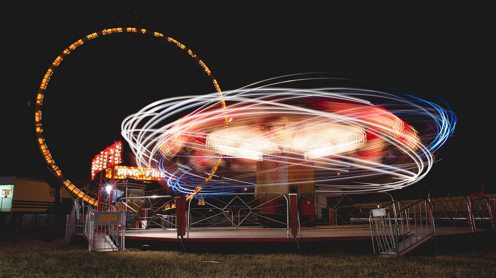 An eccentric, lively image of illuminated carnival rides creating neon light trail patterns in various colors. Original…