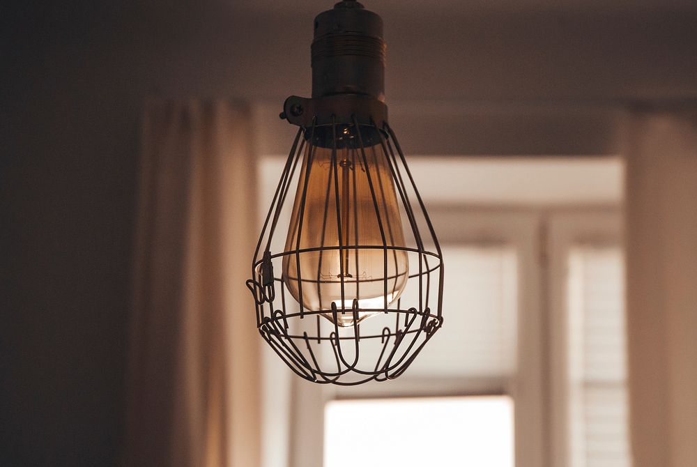 Vintage lightbulb in wire cage casing hanging from a room ceiling. Original public domain image from Wikimedia Commons