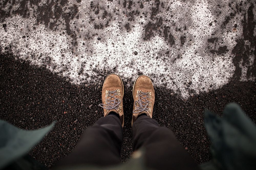 First person view of boots on the black sand by the foamy ocean in Iceland. Original public domain image from Wikimedia…