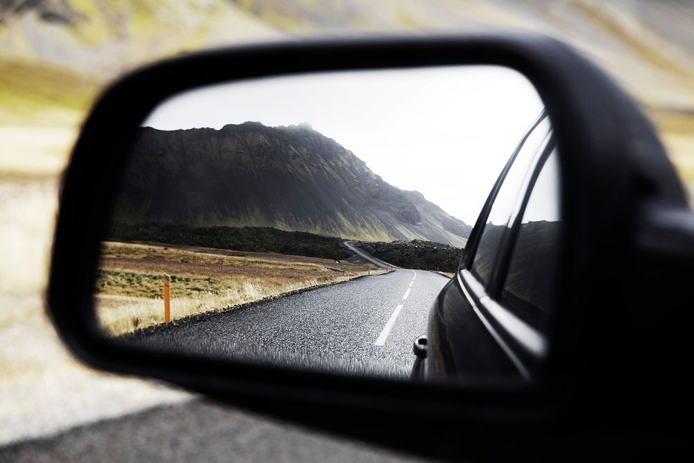 A winding highway cutting through a mountain, a dark forest and a plain field reflected in a car's side view mirror.…