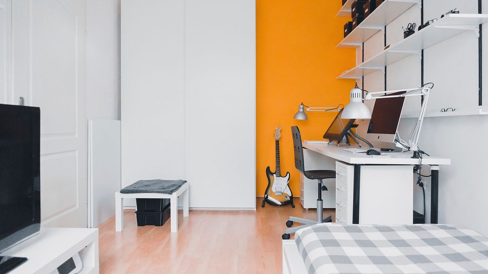 A minimally decorated room with a computer desk, a graphics tablet and a guitar against an orange wall. Original public…