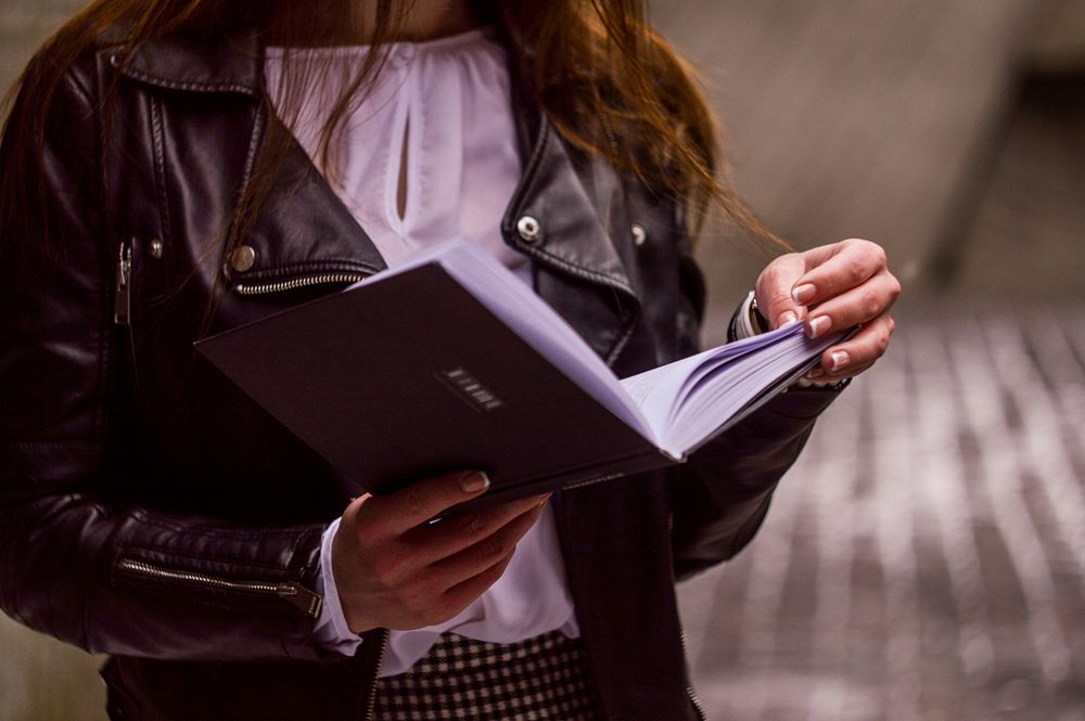 A woman in a leather jacket and white blouse holds an open book, reading. Original public domain image from Wikimedia Commons