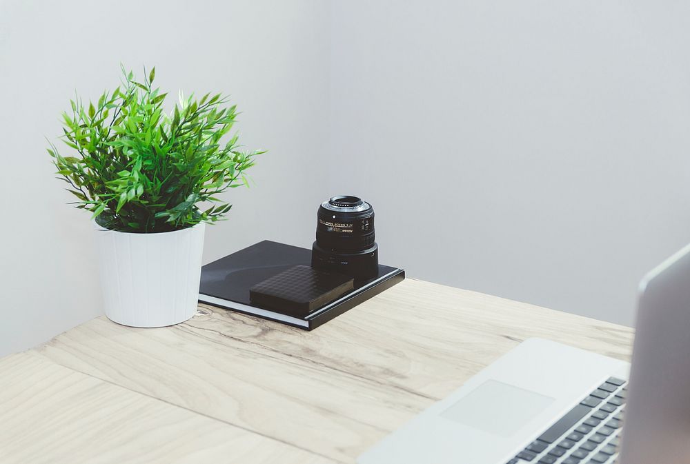 Zoom lens, a portable drive and a notebook at the edge of a table next to a potted plant. Original public domain image from…