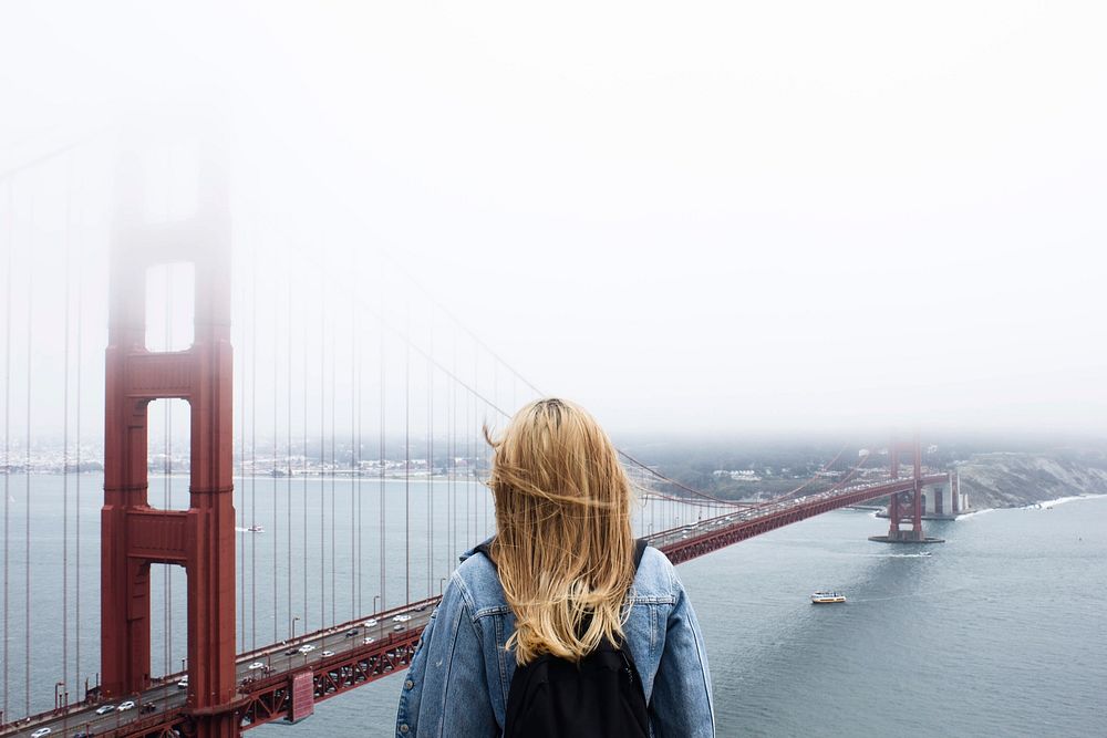 A blonde woman with a backpack looking at the Golden Gate Bridge. Original public domain image from Wikimedia Commons