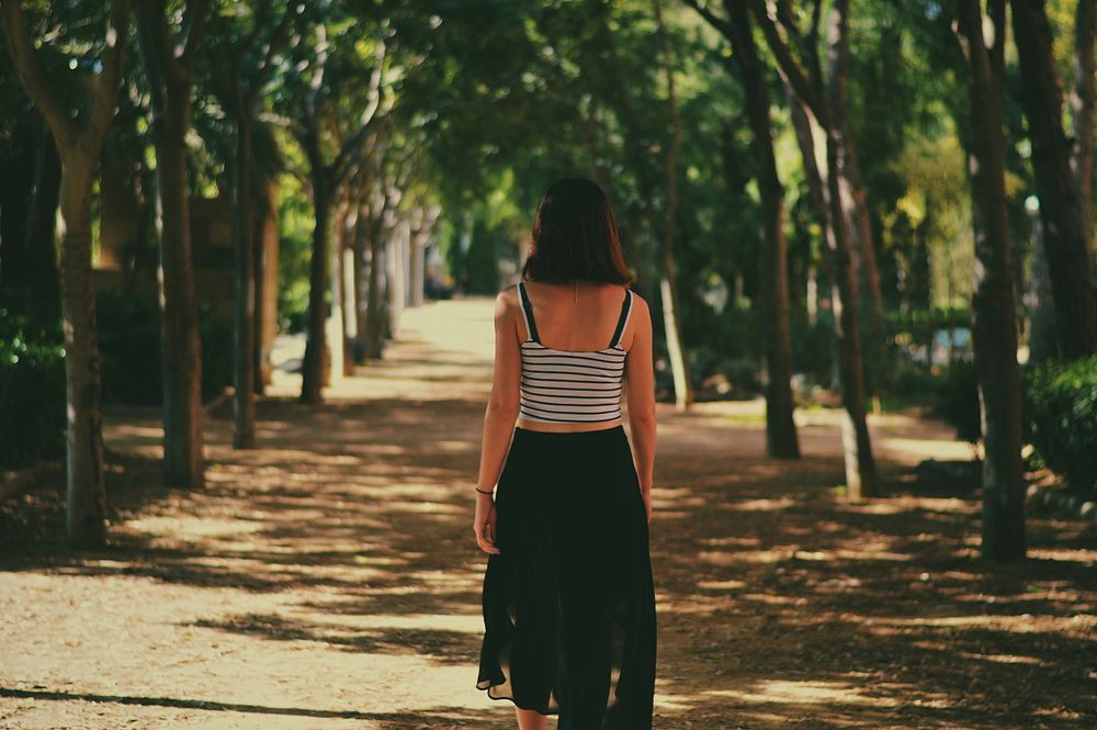 A woman in a striped tank top and black skirt walks away down a tree-lined path. Original public domain image from Wikimedia…