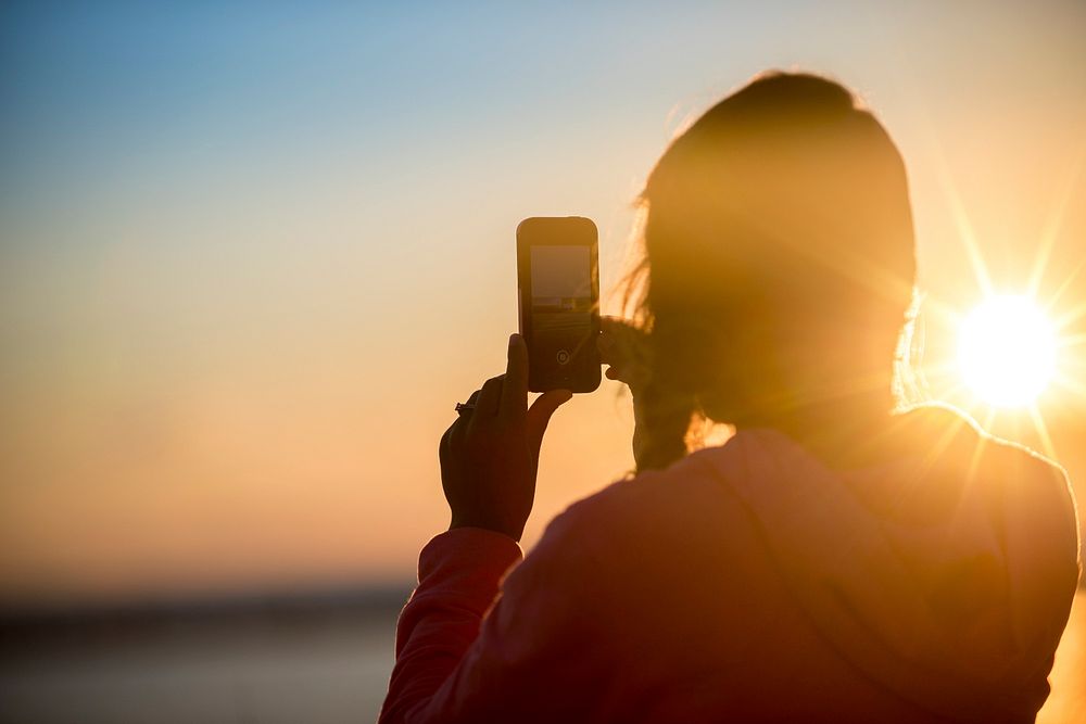 A woman on the beach taking a photo on her smartphone during golden hour. Original public domain image from Wikimedia Commons