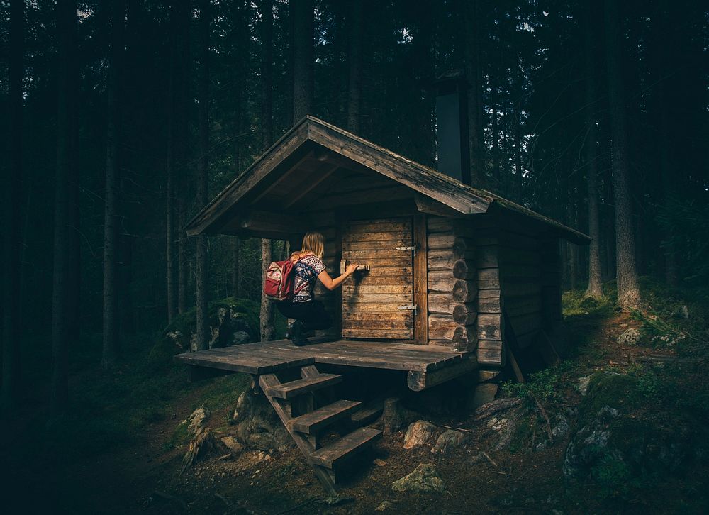 A hiker kneeling on the porch of a small wood cabin in the woods. Original public domain image from Wikimedia Commons