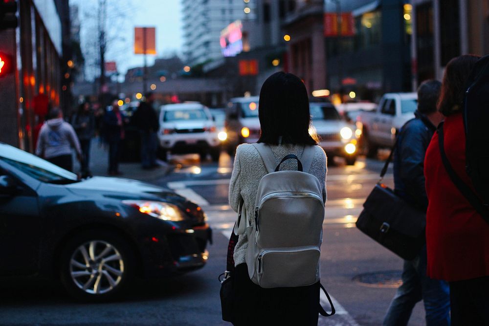 Young woman with white backpack standing on busy urban crossroads near traffic in San Francisco. Original public domain…