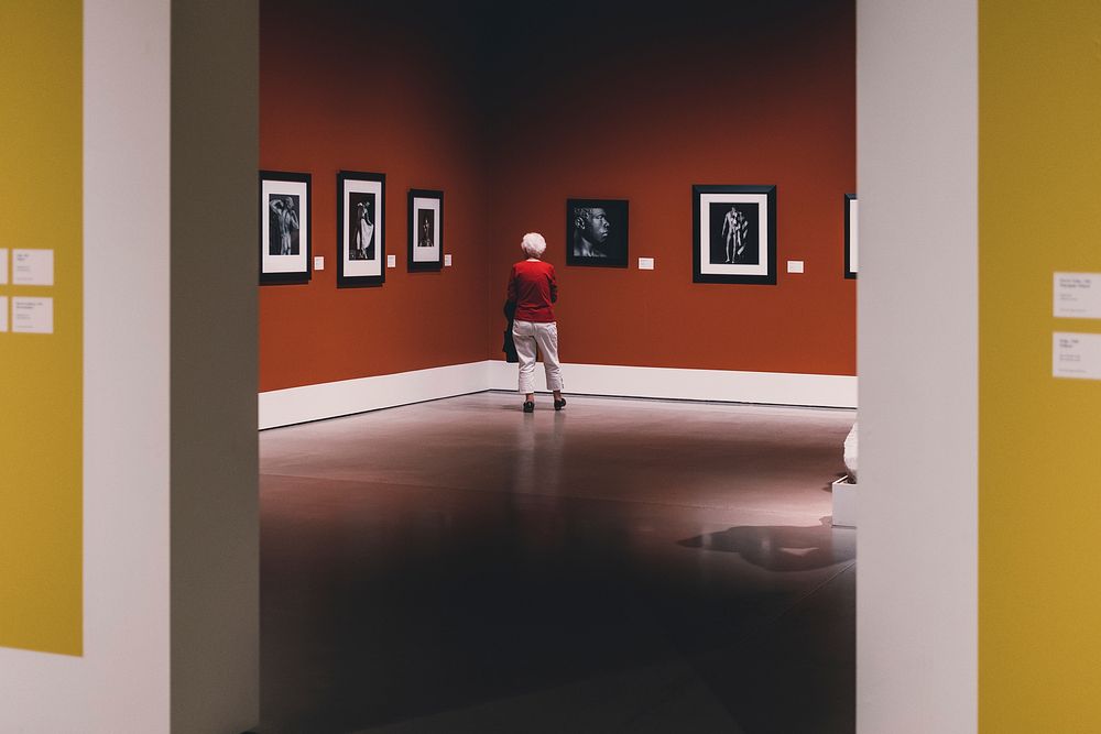 Older person in red jumper viewing artwork in gallery with marble floor and reflection. Original public domain image from…