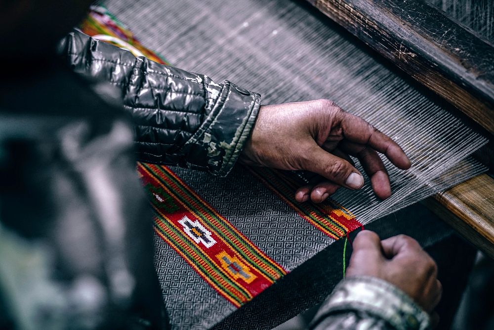 A person wearing a black quilted jacket weaving thread to create a textile on a loom in Manu Temple Road. Original public…