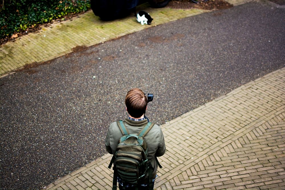 A man in a backpack holds a camera by a pathway in front of a cat. Original public domain image from Wikimedia Commons