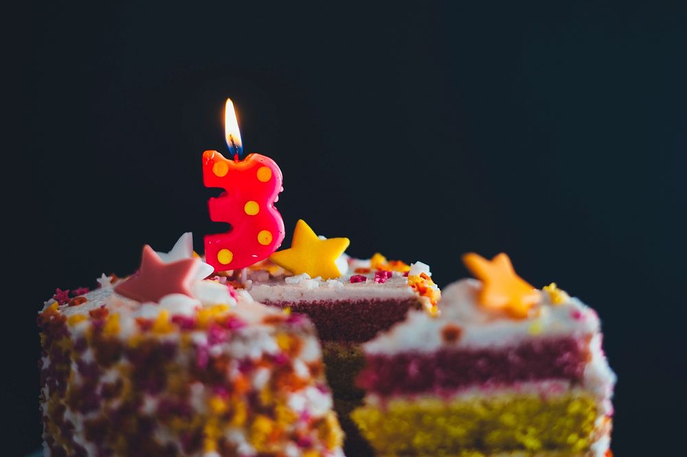 A close-up macro shot of a sliced birthday cake with a number 3 candle burning on top. Original public domain image from…